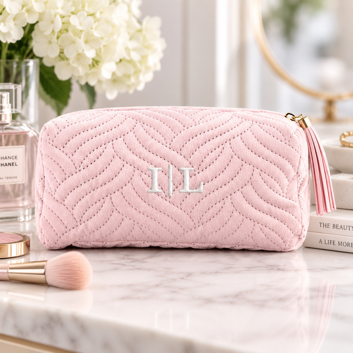 Blush pink quilted velvet makeup bag embroidered with the initials I and L, displayed on a vanity with perfume, white flowers, and a makeup brush.