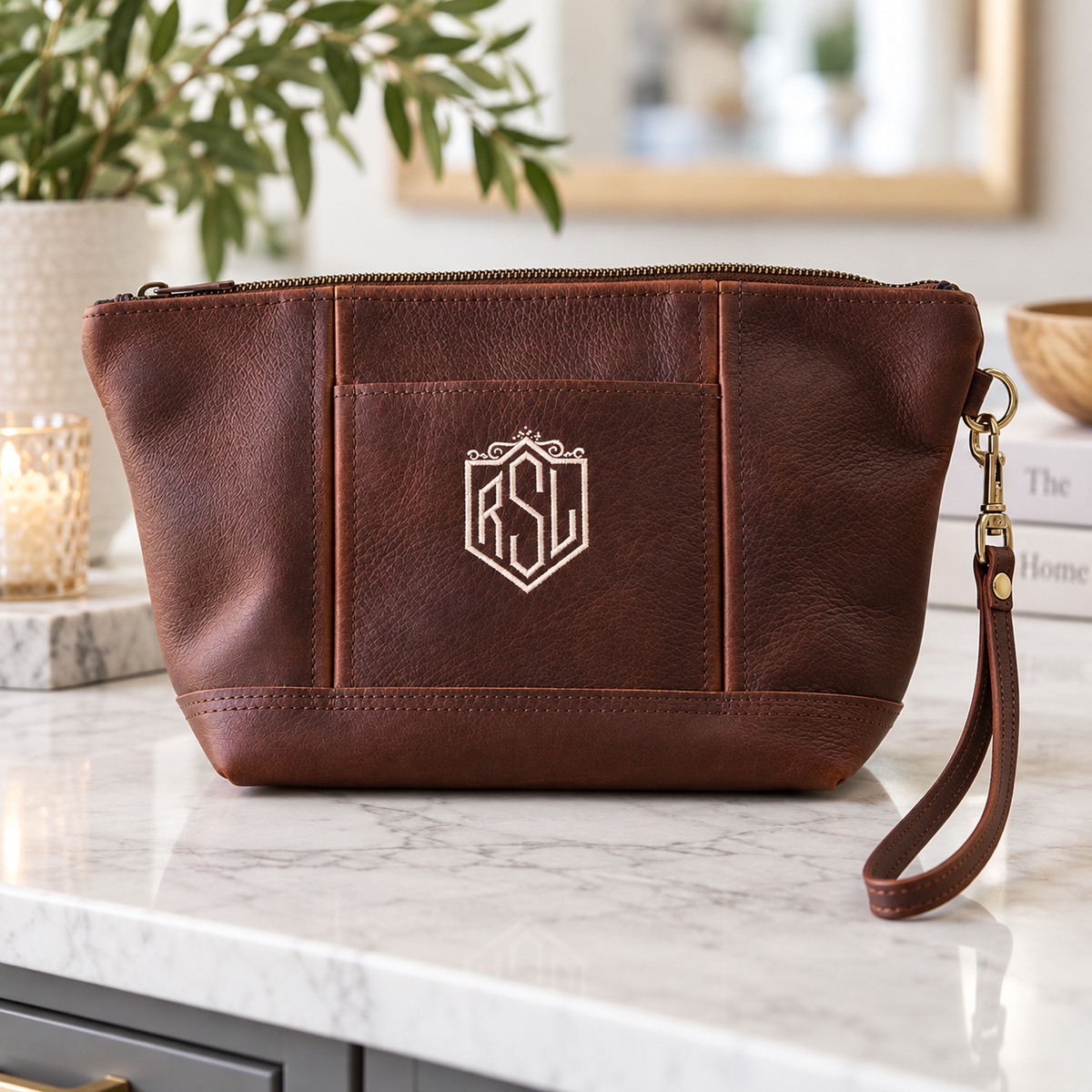 Chocolate brown leather toiletry bag with monogrammed shield logo sample, wrist strap, and brass hardware on marble countertop