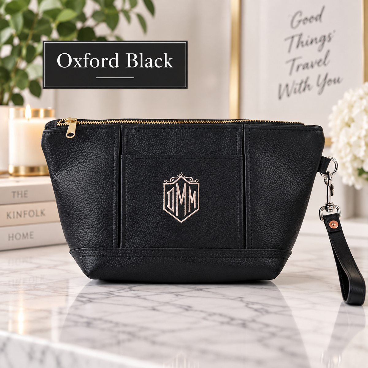 Black leather toiletry bag with sample initials "MM" engraved on front panel, gold zipper, wristlet strap, displayed on marble surface with decorative background