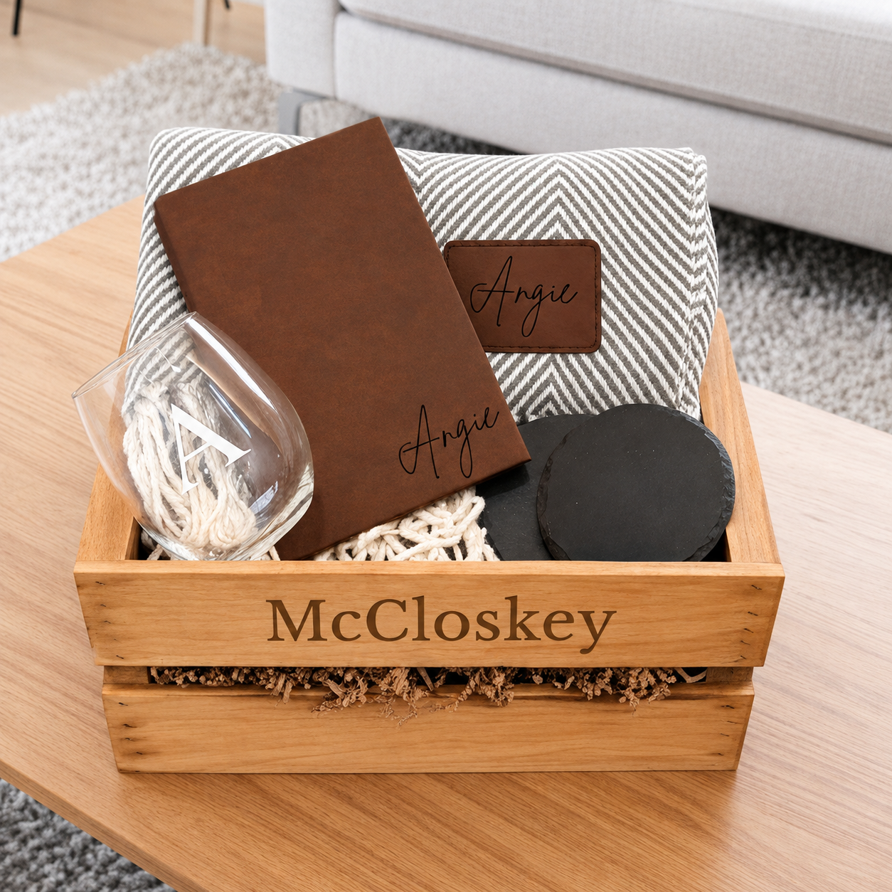 Wooden gift box with brown leather journal engraved "Angie", cozy blanket, wine glass, and black candle on wood table