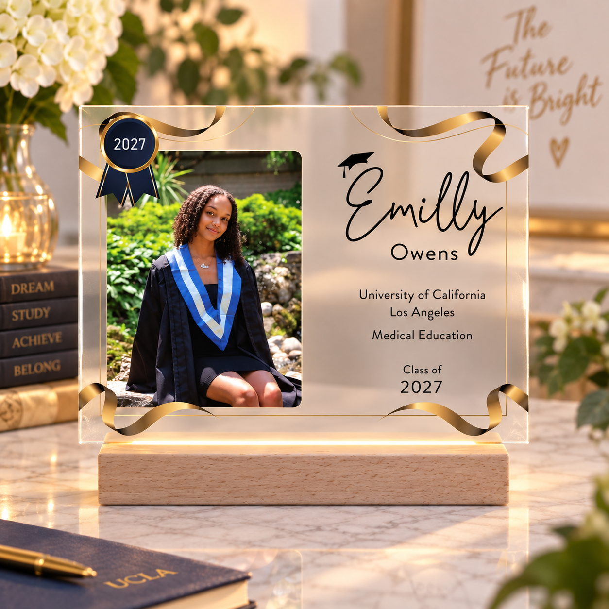Light-up graduation plaque with gold ribbon frame, 'Emilly Owens,' UCLA, and class of 2027 engraved beside her photo, glowing on its wood LED base.
