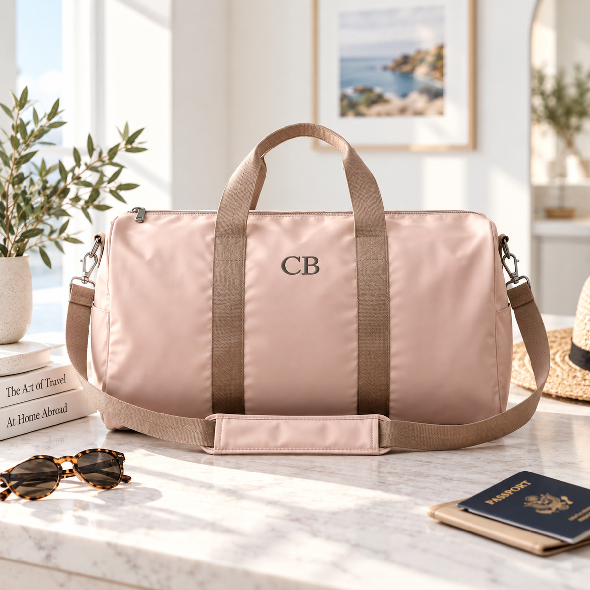 Blush pink personalized duffle bag with 'CB' embroidered on the front, styled on a marble console with sunglasses, travel books, and a passport.
