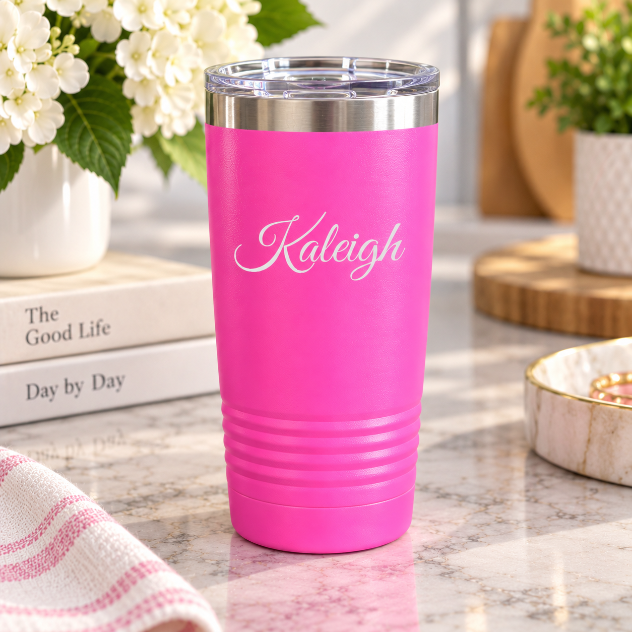 Bright pink insulated coffee tumbler engraved with the name Kaleigh in a flowing script font, on a marble counter with white flowers and books.
