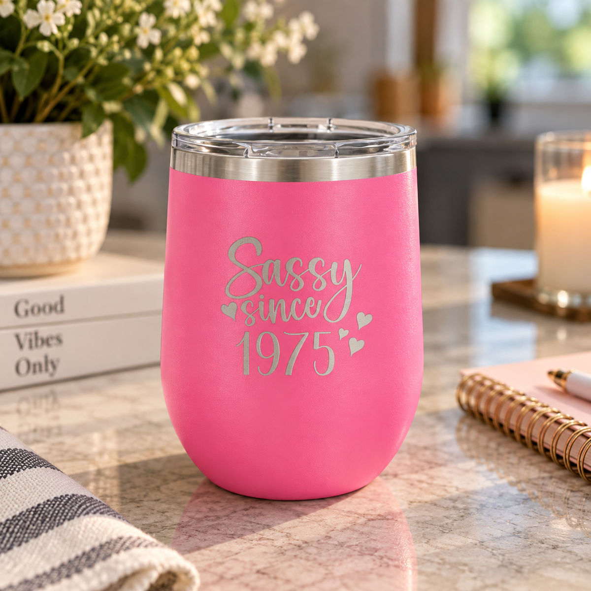 Pink stemless wine tumbler laser engraved with a 'Sassy since 1975' 50th birthday design, on a desk with books and a candle.