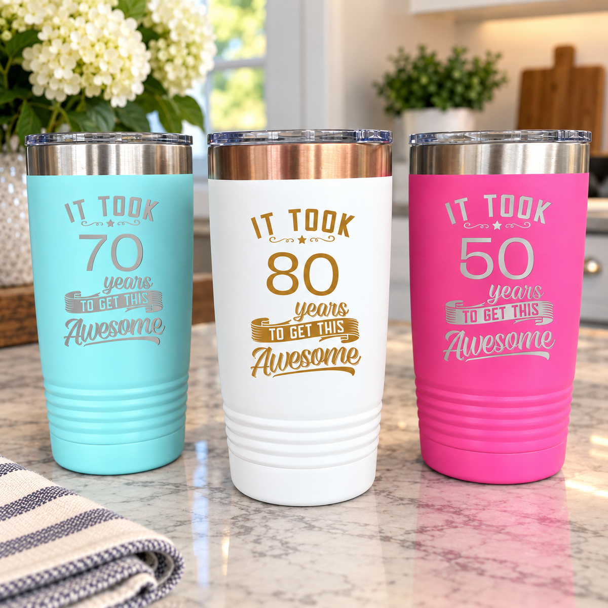 Three engraved stainless steel tumblers in turquoise, white and pink showing sample age milestone designs at 70, 80 and 50 years, displayed on marble.