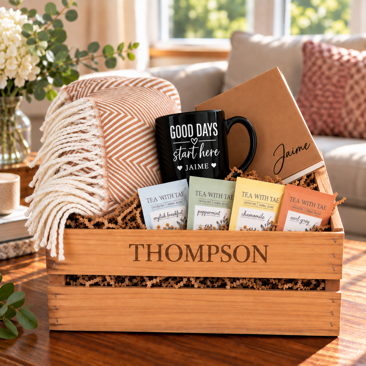 Wooden crate personalized with a family name, filled with a khaki throw blanket, engraved mug, leatherette journal, and gourmet tea sachets on a coffee table