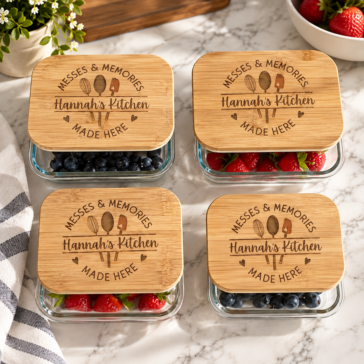 Four glass food storage containers with engraved bamboo lids reading Hannah's Kitchen, filled with blueberries and strawberries on a marble counter