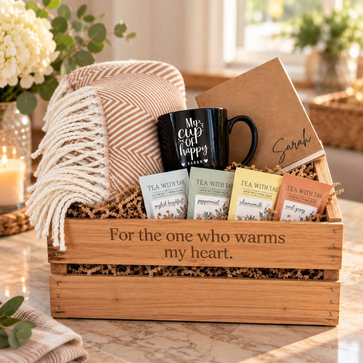 Cedar gift crate engraved with a heart-warming message, holding a black personalized mug, cream throw blanket, tan journal, and four tea sachets on a marble counter