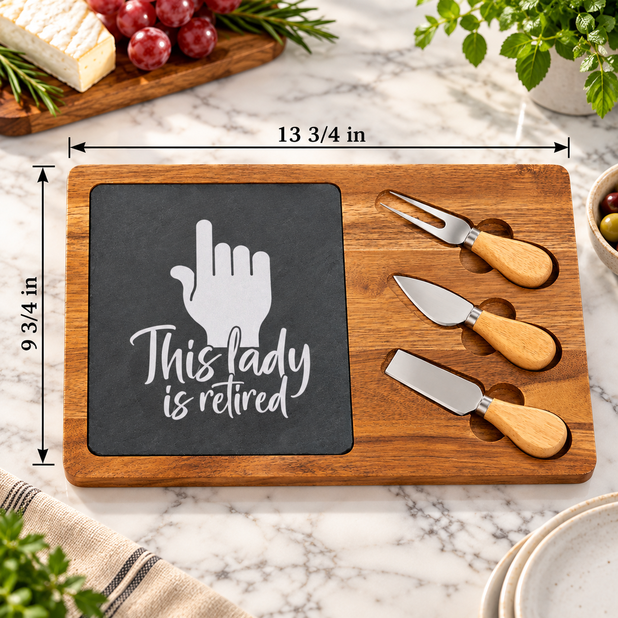 Retirement serving board measuring 9 3/4 by 13 3/4 inches, with a wood base, an engraved slate insert reading This Lady Is Retired, and three serving tools.
