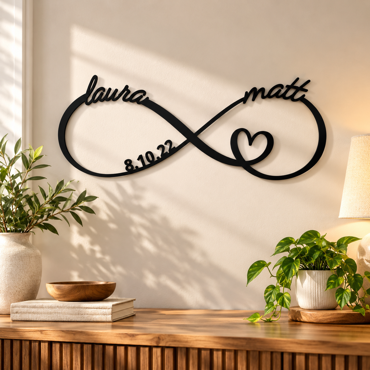 Black metal infinity sign mounted above a wooden console table, personalized with names laura and matt, a heart, and the date 8.10.22, styled with plants and a table lamp