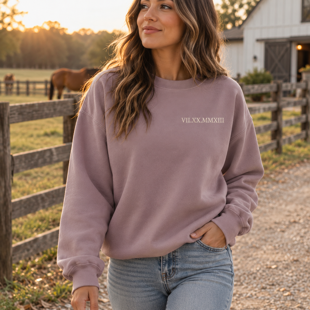 Woman wearing a lilac purple embroidered sweatshirt walking along a wooden fence on a farm at sunset, with a horse grazing in the background