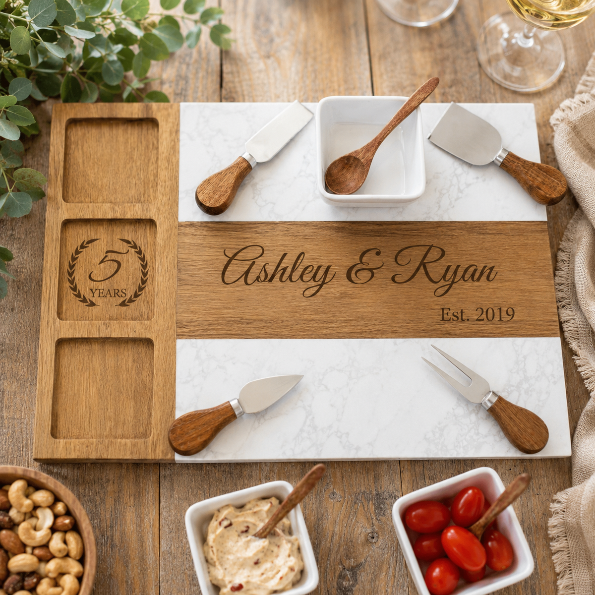 Marble and acacia wood anniversary charcuterie board engraved with a couple's names and wedding year, set with cheese knives, wine, and small bowls of nuts and tomatoes.