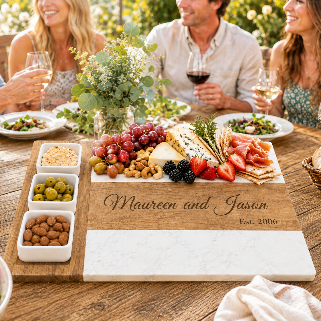 Personalized anniversary charcuterie board engraved with a couple's names, held at an outdoor dinner table while friends toast with wine around cheese, charcuterie, and fruit.