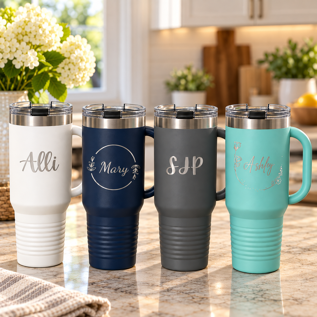 Four personalized tumblers with handles on a kitchen counter, in white, navy, grey, and teal, each engraved with a different name or initials and design.