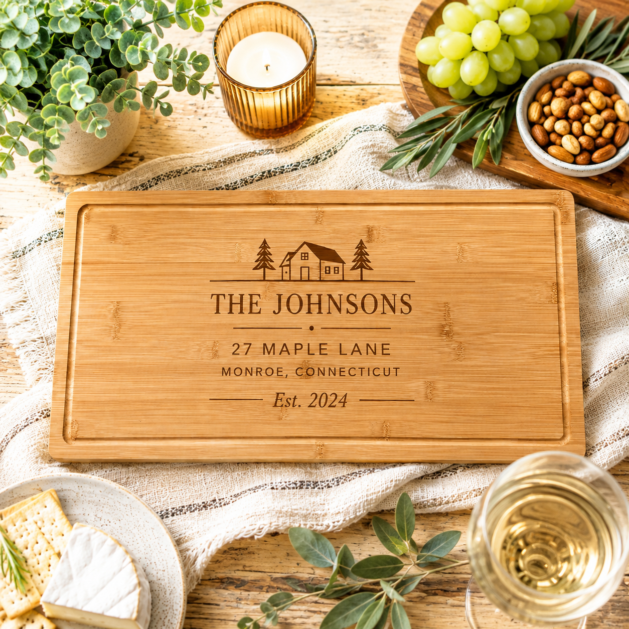 Personalized bamboo closing gift board engraved 'The Johnsons, 27 Maple Lane, Monroe, Connecticut, Est. 2024' on a wooden table with grapes, almonds and candlelight.