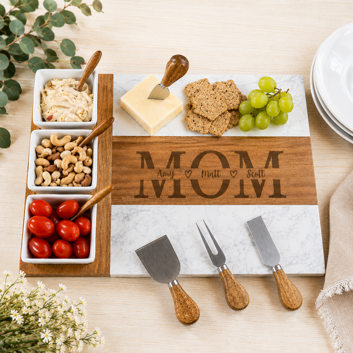A wooden and marble cheese board set arranged on a light wood table, featuring a rectangular board engraved with the name 'MOM' and three small white serving dishes with cheeses, crackers, nuts, grapes, and cherry tomatoes. Three small hand tools with wooden handles are placed in front.