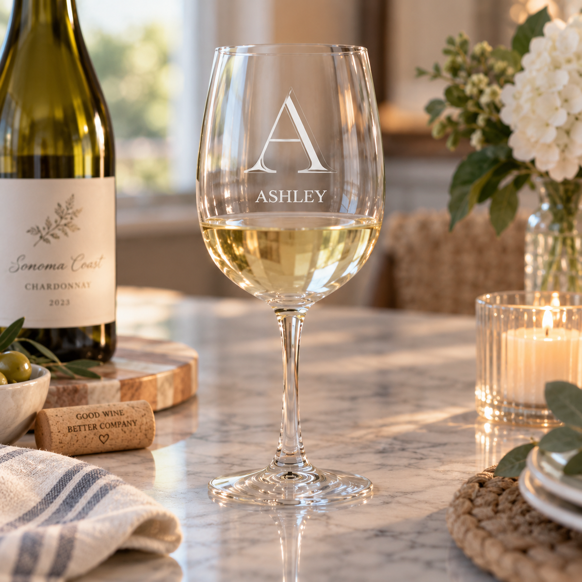 A personalized wine glass with the name 'ASHLEY' and monogram 'A' engraved, filled with white wine on a marble table next to a wine bottle and candle.