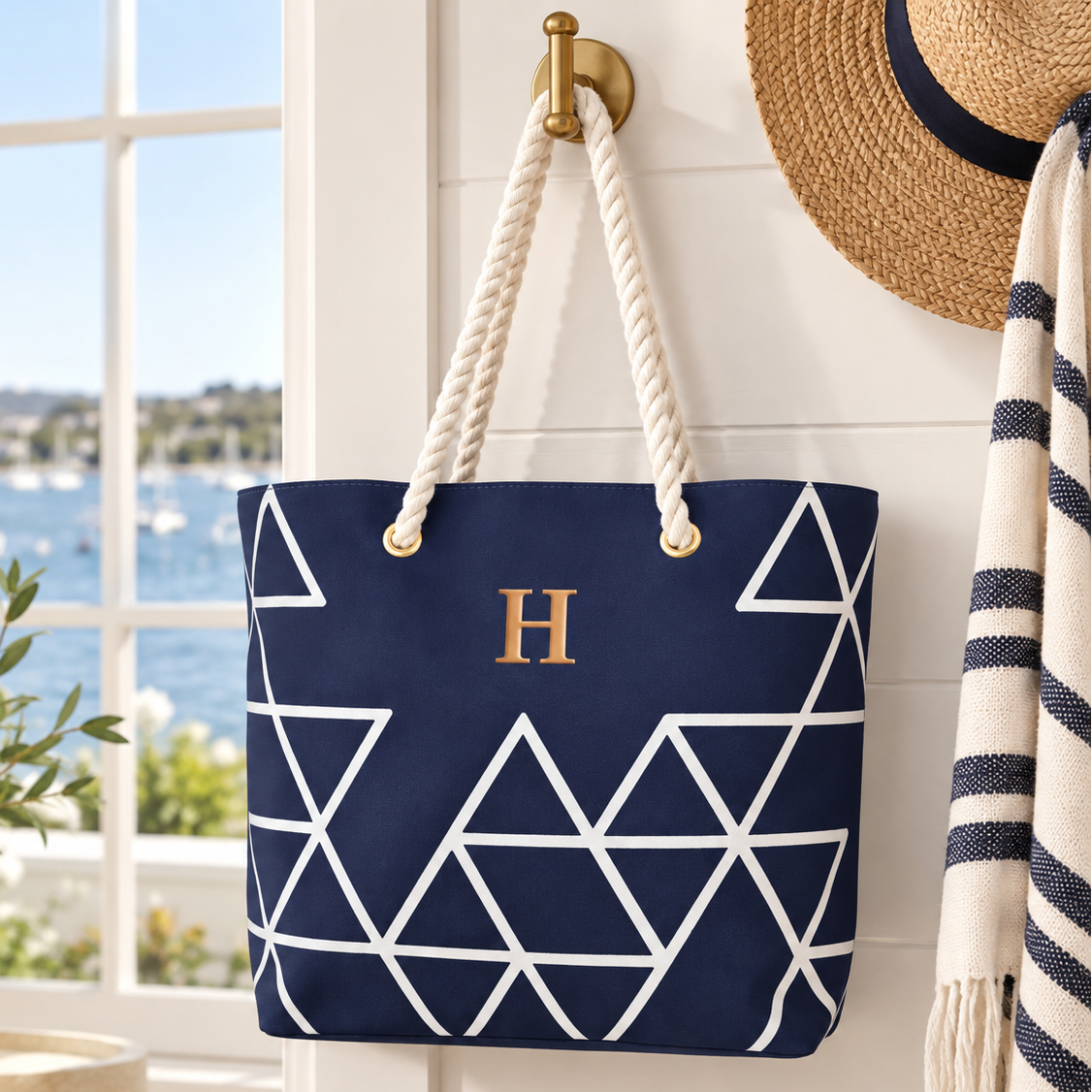 A navy blue canvas tote bag with a white geometric triangle pattern and a gold monogram letter 'H' on the front, hanging from a brass hook on a white paneled wall. The background shows a bright window with a coastal view, a straw hat, and a striped throw blanket.