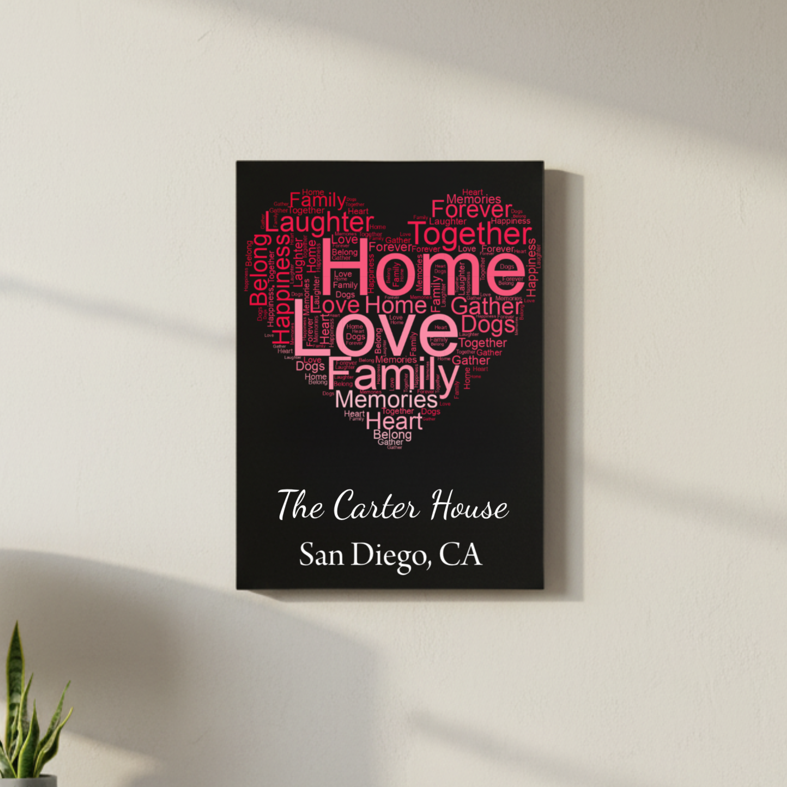Black home decor sign with heart-shaped word cloud in pink and gray text reading "home," "love," "family," "memories," and "together," personalized with sample text "The Carter House" and "San Diego, CA" below in white script and serif fonts.