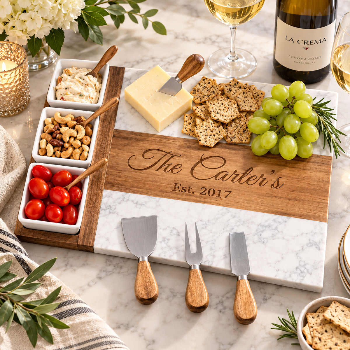 Personalized wooden and marble charcuterie board set with three cheese knives, styled with crackers, grapes, tomatoes, and wine on a marble countertop.