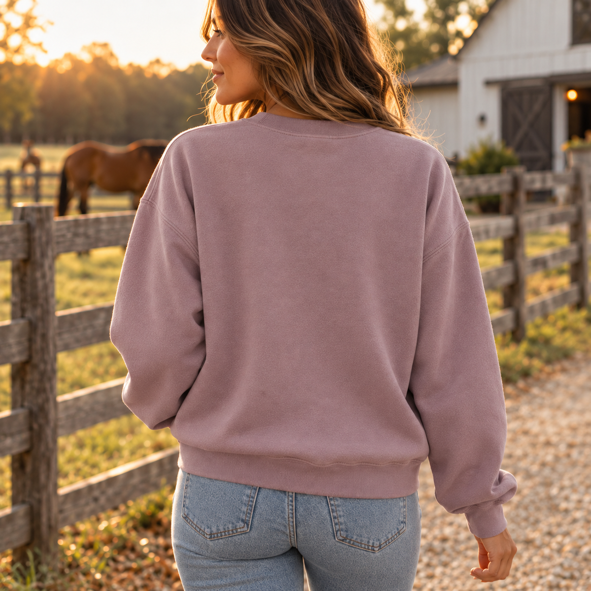 A woman viewed from behind wearing a dusty mauve crewneck sweatshirt and light blue jeans, standing on a gravel path next to a wooden fence with a horse and a white barn in the background during golden hour.