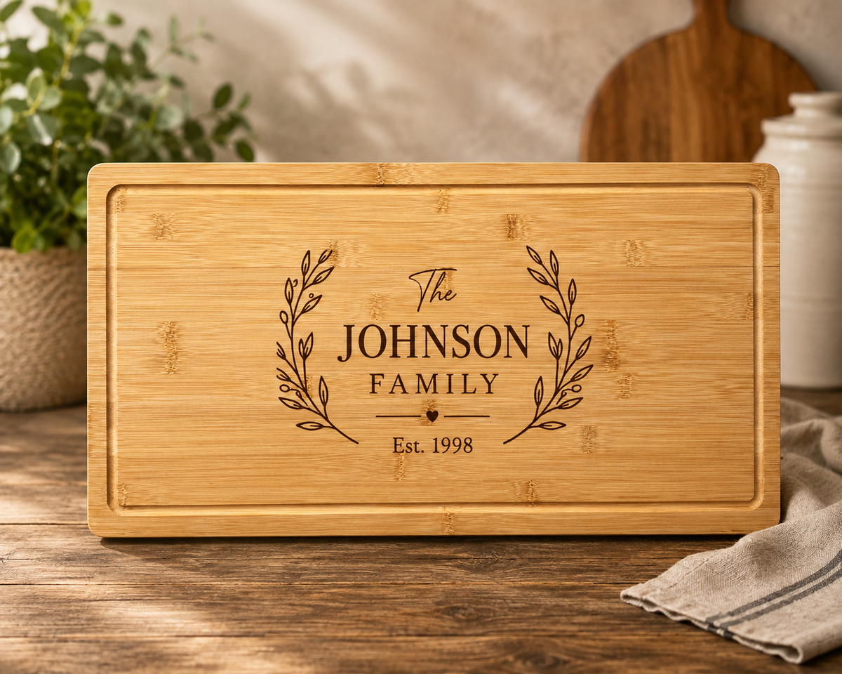 A rectangular bamboo cutting board with rounded corners, featuring a personalized engraving that reads 'The JOHNSON FAMILY Est. 1998' with leafy branches on either side.