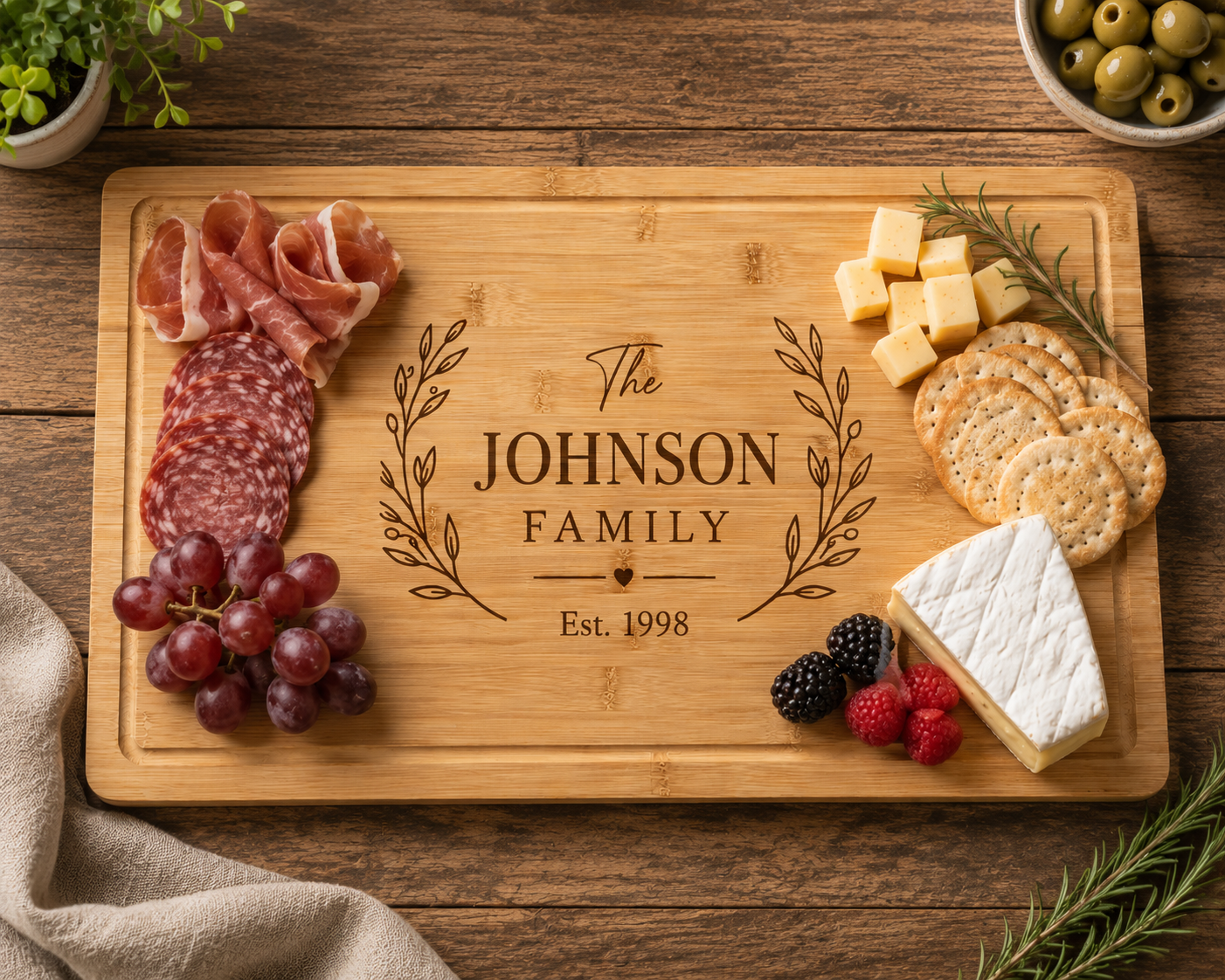 A rectangular wooden charcuterie board engraved with 'The JOHNSON FAMILY Est. 1998' and leafy flourishes, styled on a rustic wooden table with sliced salami, prosciutto, grapes, crackers, cheese, olives, and herbs.