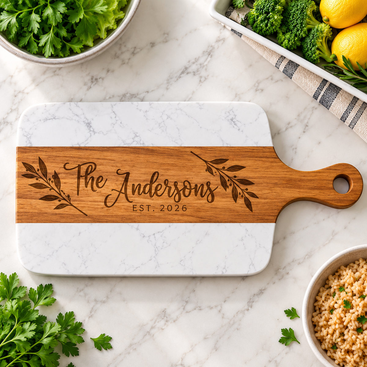 A personalized wooden cutting board with a white marble base, featuring the engraved text 'The Andersons EST. 2026' and leafy branch designs.