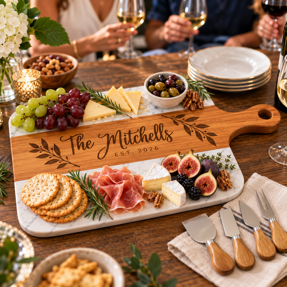 A personalized wooden charcuterie board with a white marble base, engraved with 'The Mitchells Est. 2026', styled on a wooden dining table with cheese, crackers, grapes, figs, olives, and wine glasses in the background.