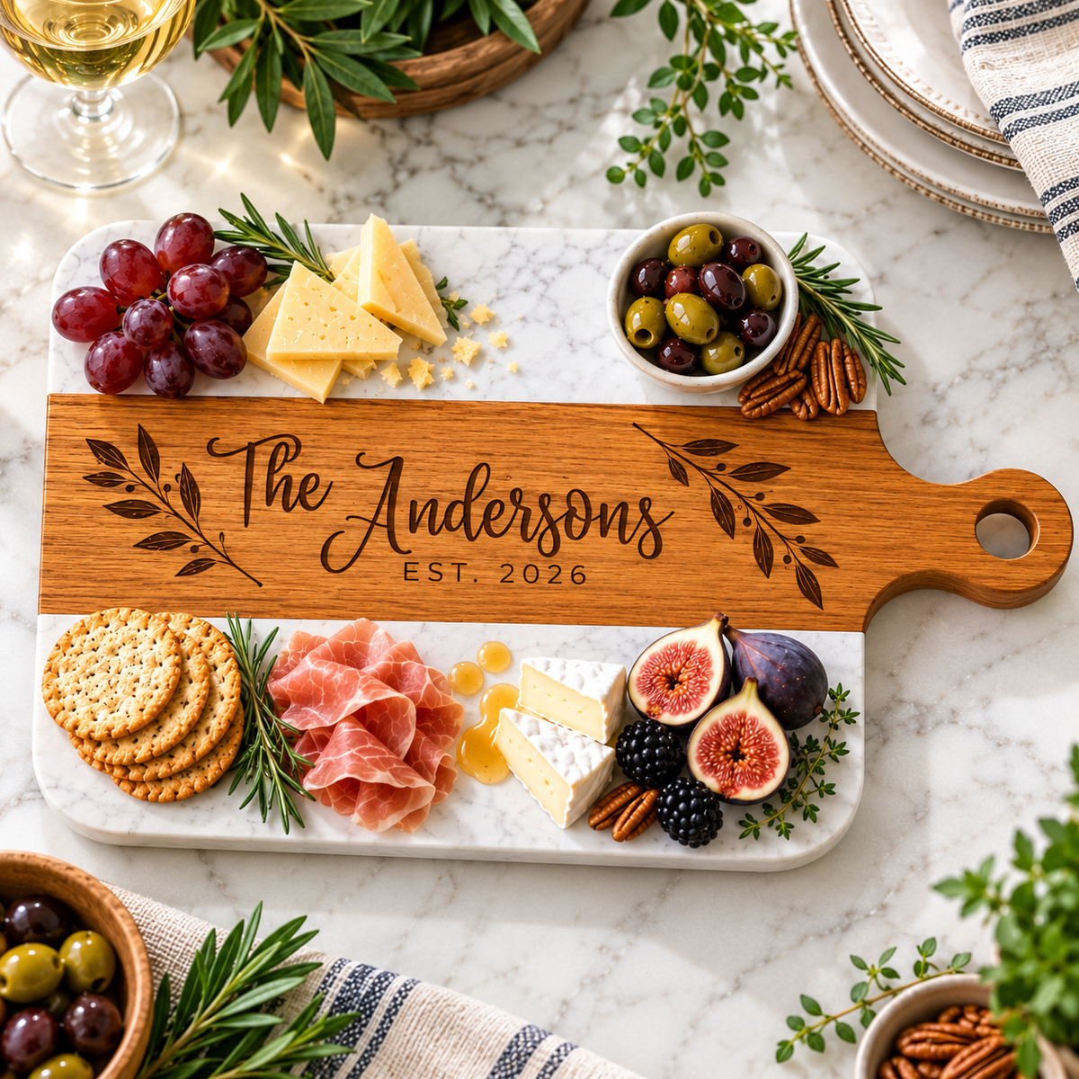 A personalized wooden charcuterie board with a white marble base, styled with cheese, crackers, grapes, olives, prosciutto, figs, and herbs on a marble countertop.