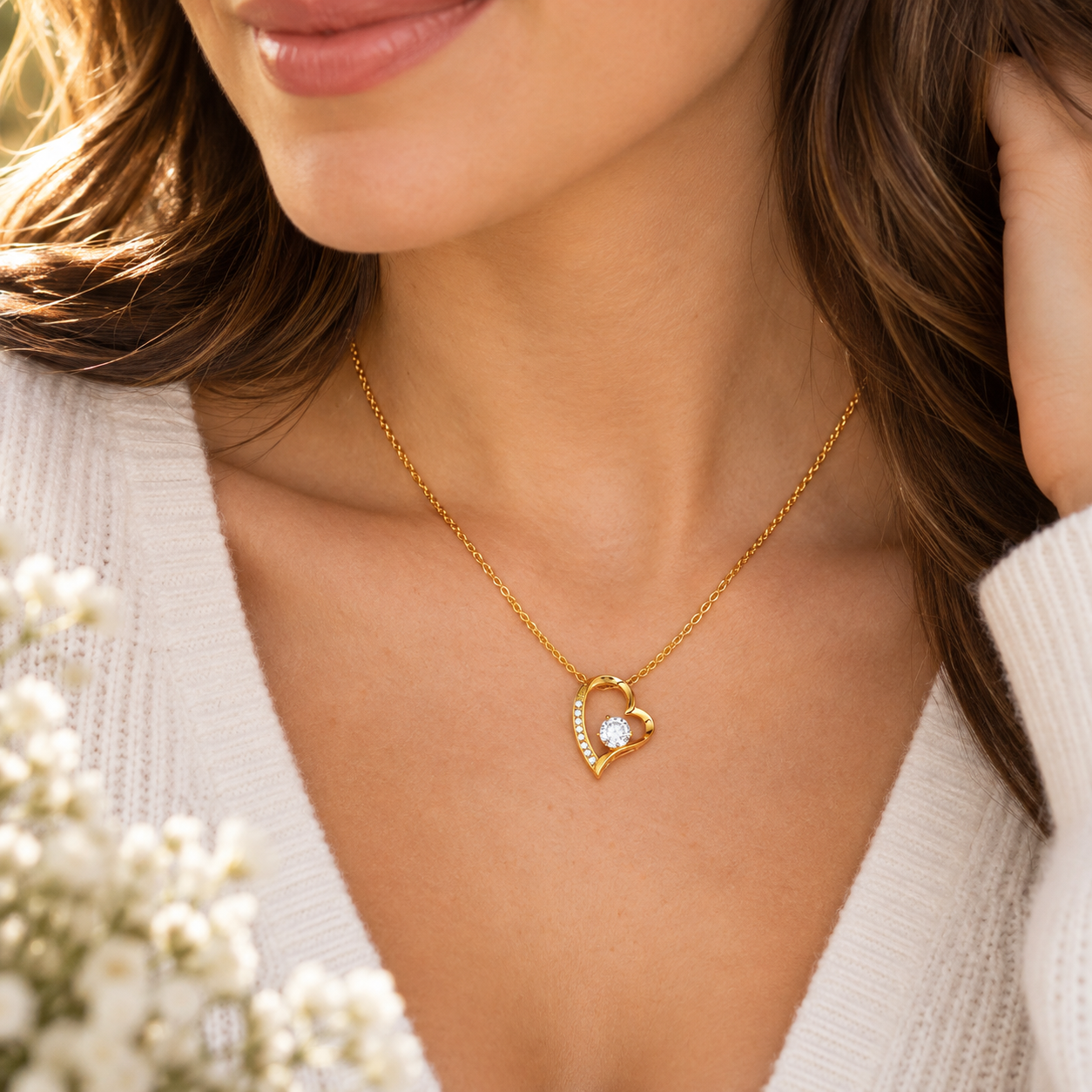 A close-up lifestyle shot of a woman wearing a delicate gold heart-shaped pendant necklace with a clear central gemstone, styled with a white knit top and soft white flowers in the foreground.