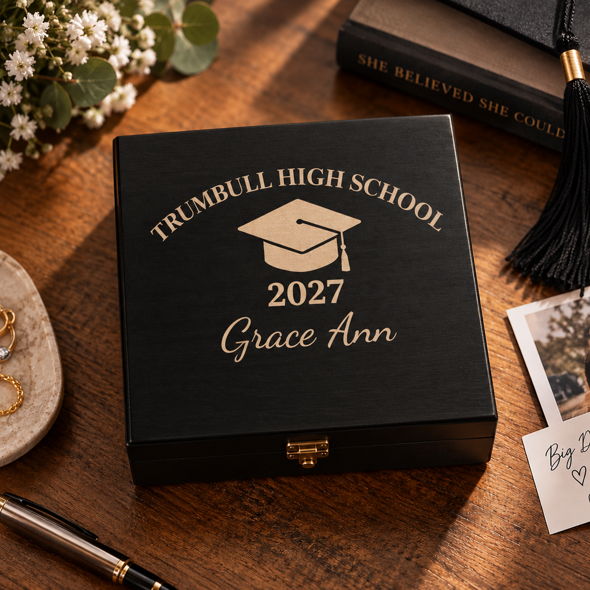 Black wooden graduation keepsake box engraved in gold with Trumbull High School, a graduation cap, 2027 and Grace Ann, on a desk beside a graduation tassel