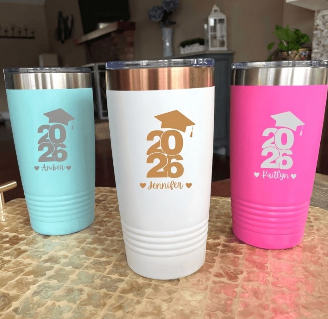 Three insulated tumblers in blue, white, and pink with '2026' graduation gift design on a countertop.