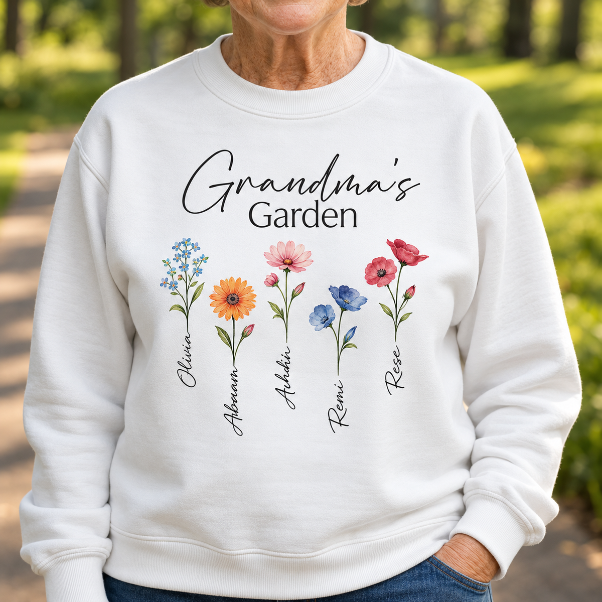 White crewneck sweatshirt with printed text "Grandma's Garden" and five watercolor flowers, each labeled with sample names Olivia, Aiden, Stella, Rosie, and Rae