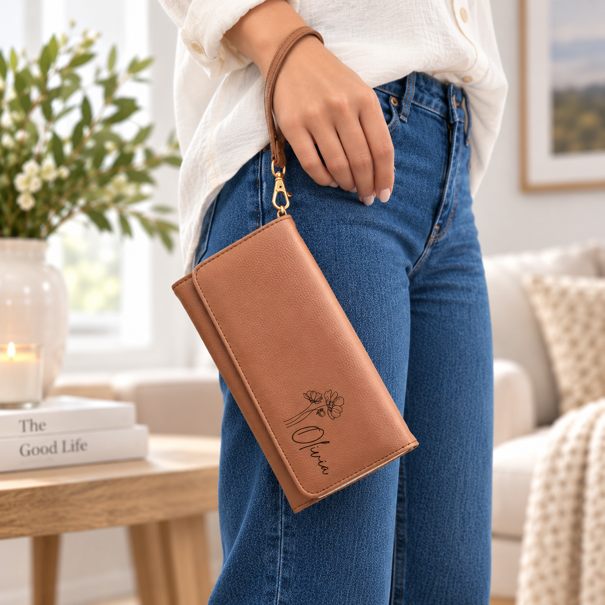 A woman in a white blouse and blue jeans holds a tan faux-leather wristlet clutch with a gold-tone clasp and floral monogram. The clutch has a matching wrist strap and is shown in a bright, modern living room setting.