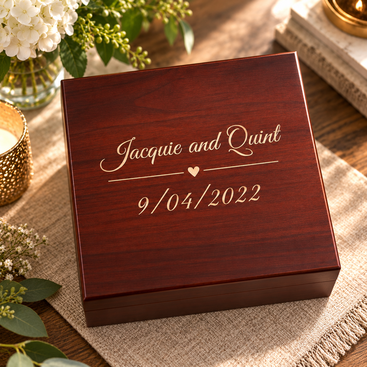 A personalized dark reddish-brown wooden keepsake box with gold engraving reading 'Jacquie and Quint' and the date '9/04/2022' with a small heart, styled on a burlap runner with flowers and candles in a warm setting.