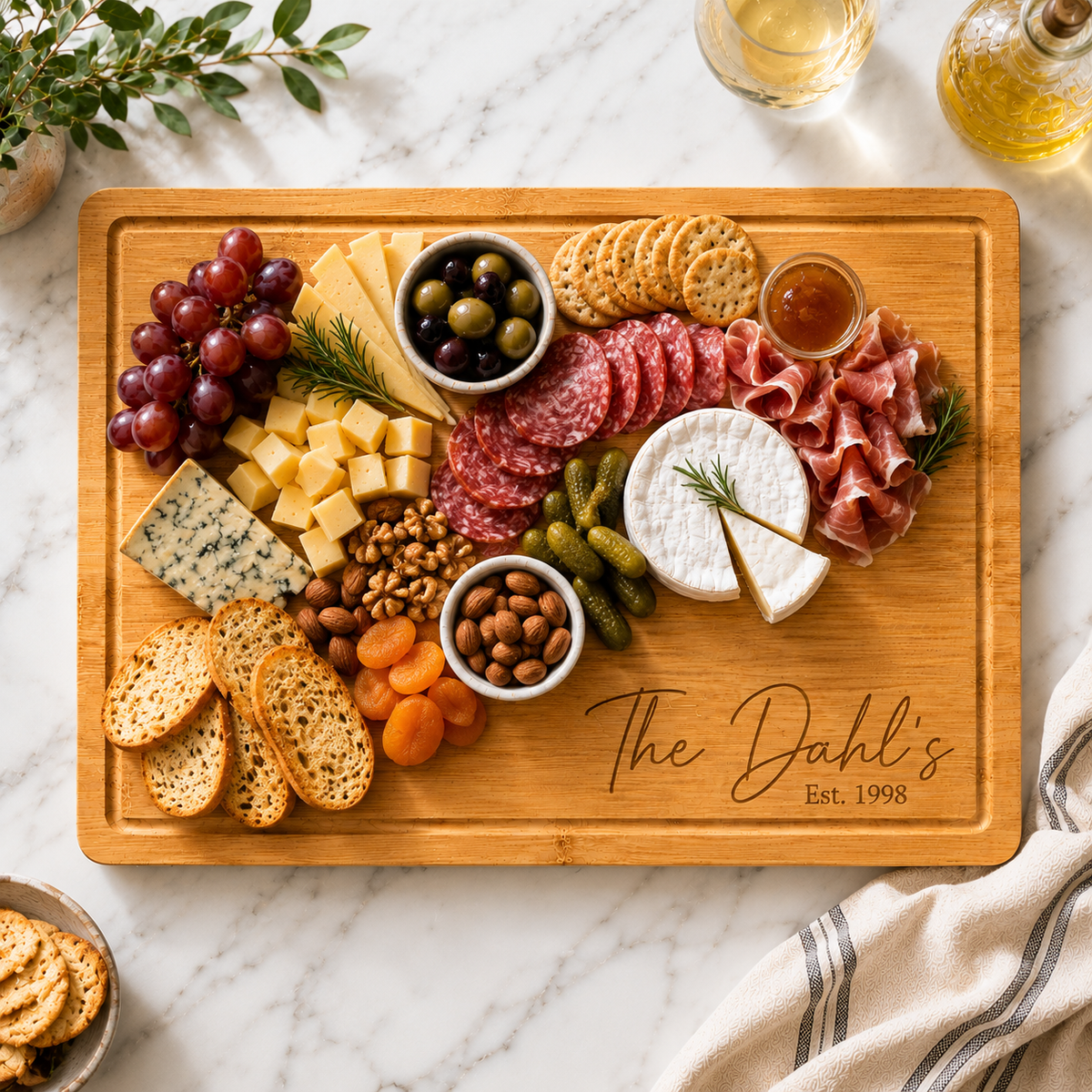 A wooden charcuterie board with an engraved script reading 'The Dahl's Est. 1998', filled with cheeses, cured meats, crackers, grapes, olives, pickles, nuts, and dried fruit on a white marble surface.