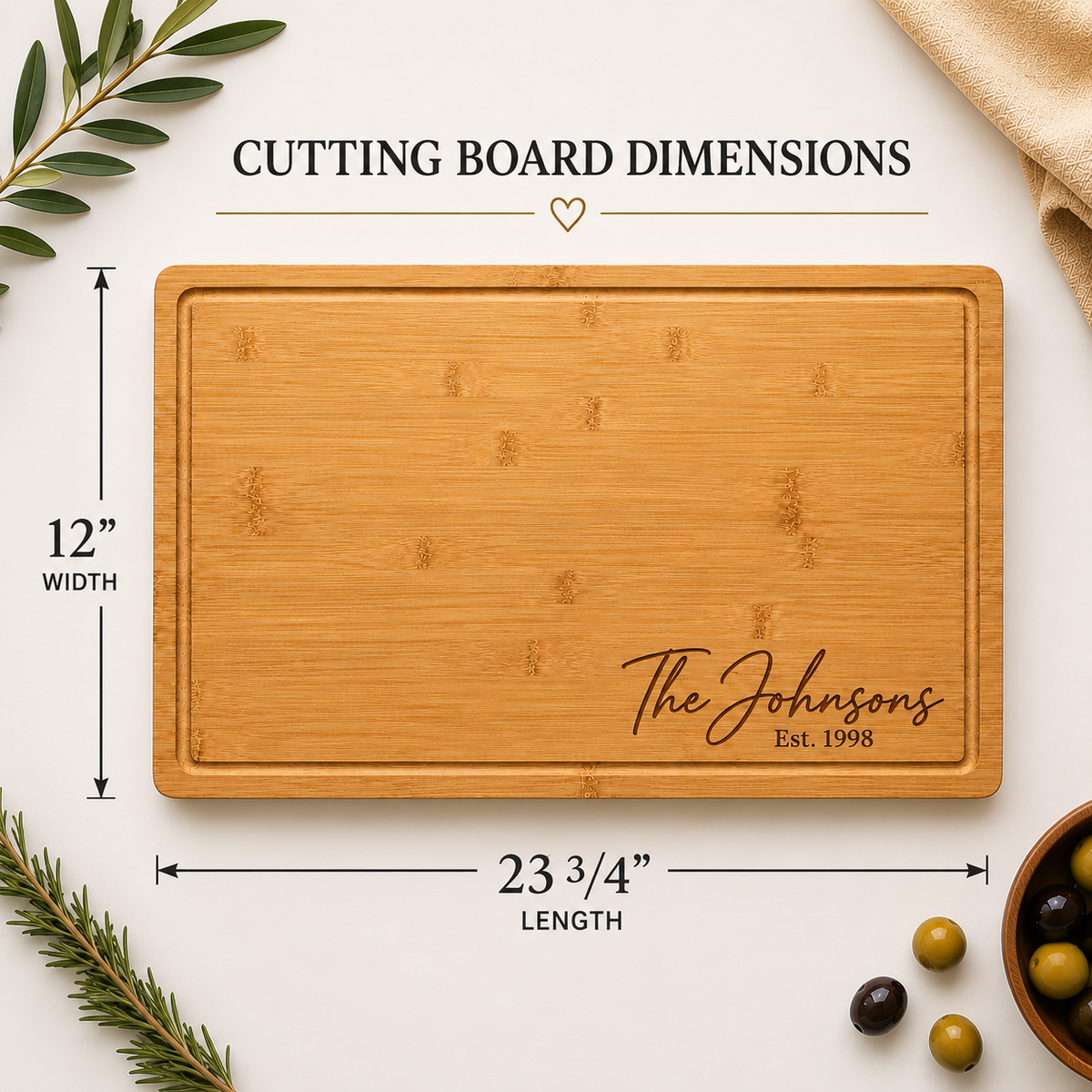 A rectangular bamboo cutting board with rounded corners and engraved script reading 'The Johnsons Est. 1998,' shown on a light neutral background with dimension callouts indicating 12 inches wide by 23 3/4 inches long.