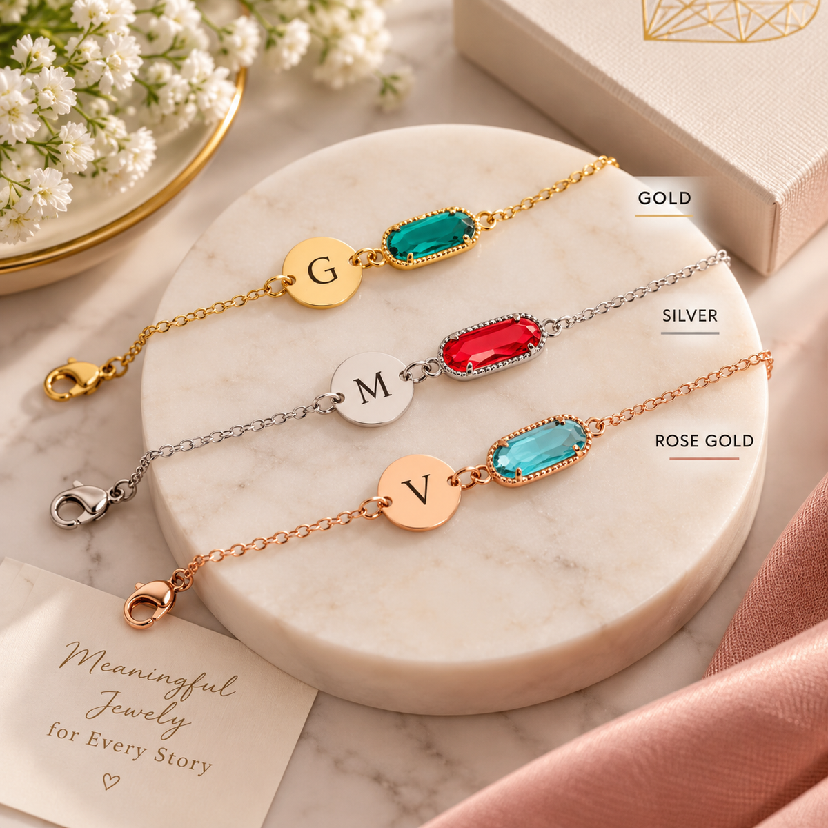 Three personalized initial bracelets with rectangular gemstones are arranged on a round white marble tray. The bracelets are labeled Gold, Silver, and Rose Gold, with the letters G, M, and V engraved on gold, silver, and rose gold discs respectively.