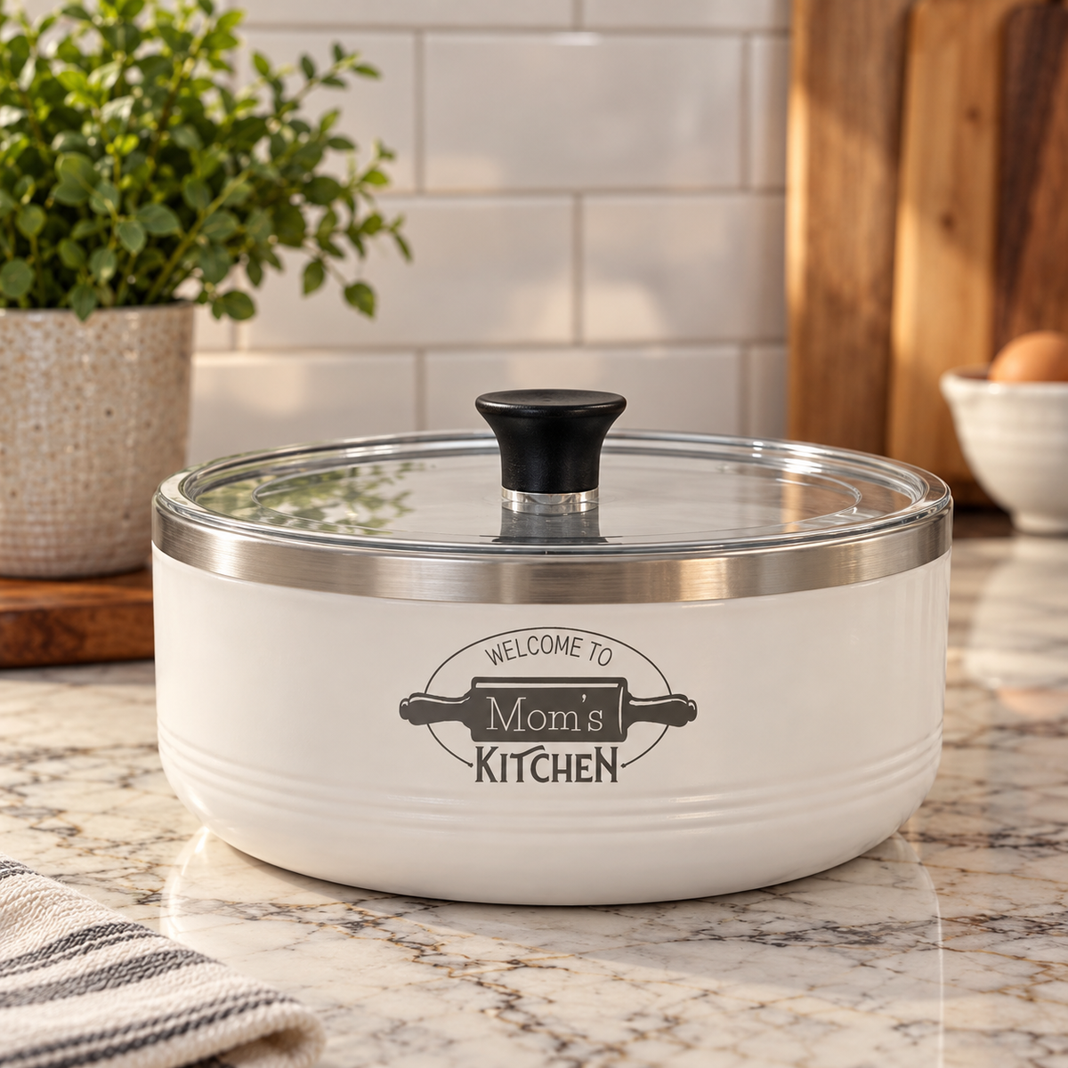 A white ceramic cooking pot with a clear glass lid and black handle, featuring a custom 'Mom's Kitchen' graphic on the front, set on a kitchen countertop.