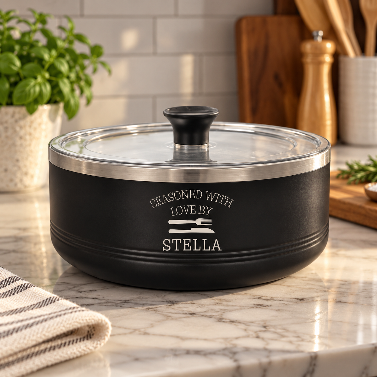 A black ceramic cooking pot with a clear glass lid and black handle, featuring the white printed text 'SEASONED WITH LOVE BY STELLA' and a fork and knife graphic, sitting on a marble kitchen countertop.