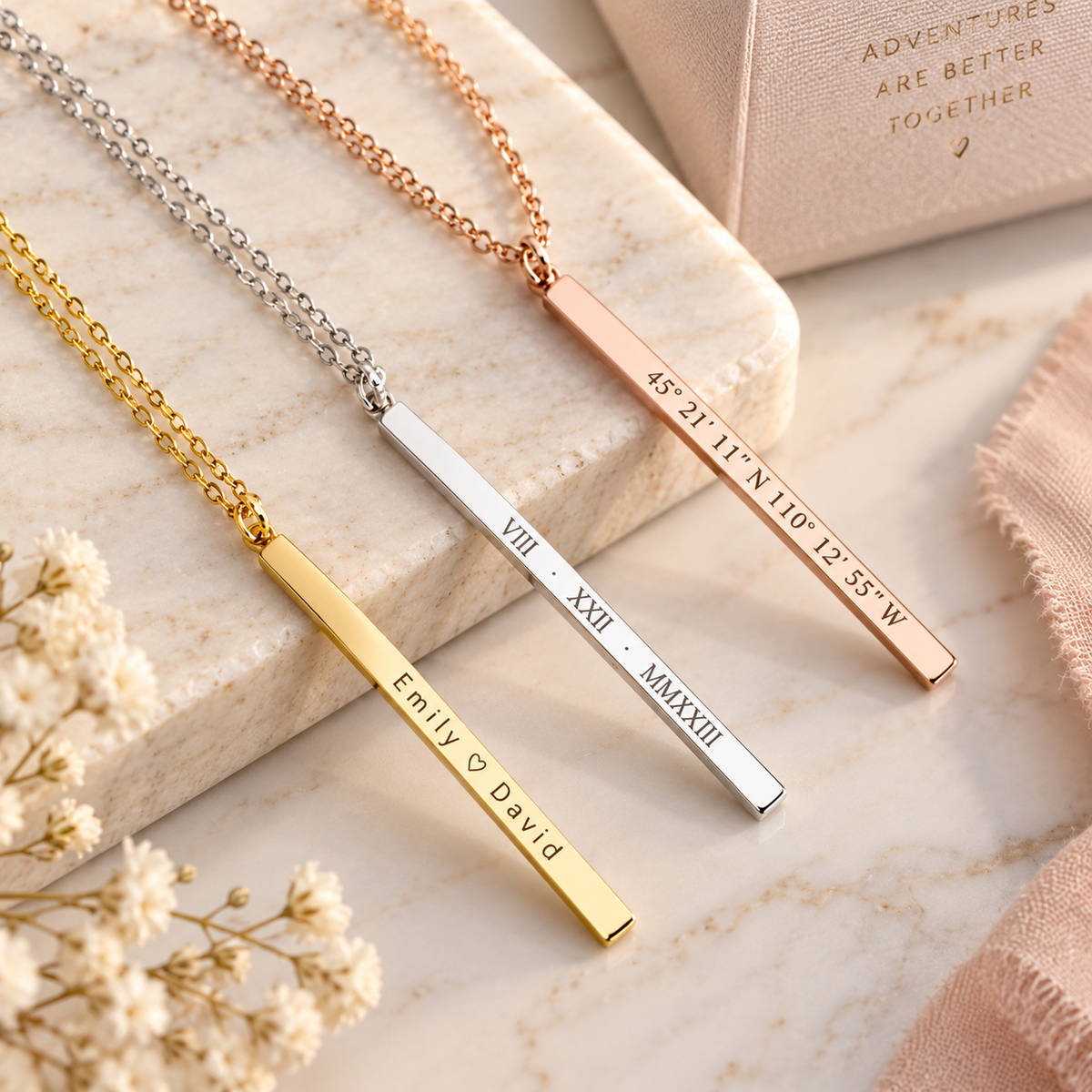 Three personalized coordinate necklaces in gold, silver, and rose gold with engraved sample coordinates and names displayed on marble surface.