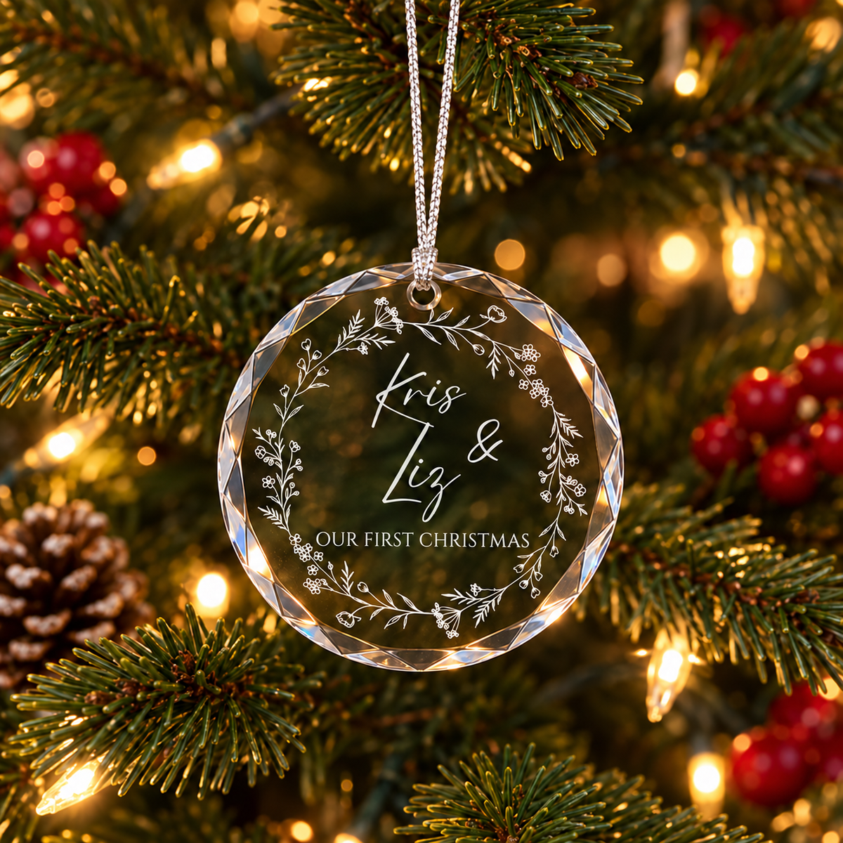 A round clear acrylic Christmas ornament engraved with a floral wreath and the names 'Kris &amp; Liz' and 'OUR FIRST CHRISTMAS,' hanging from a silver string on a decorated Christmas tree with warm lights and pinecones.