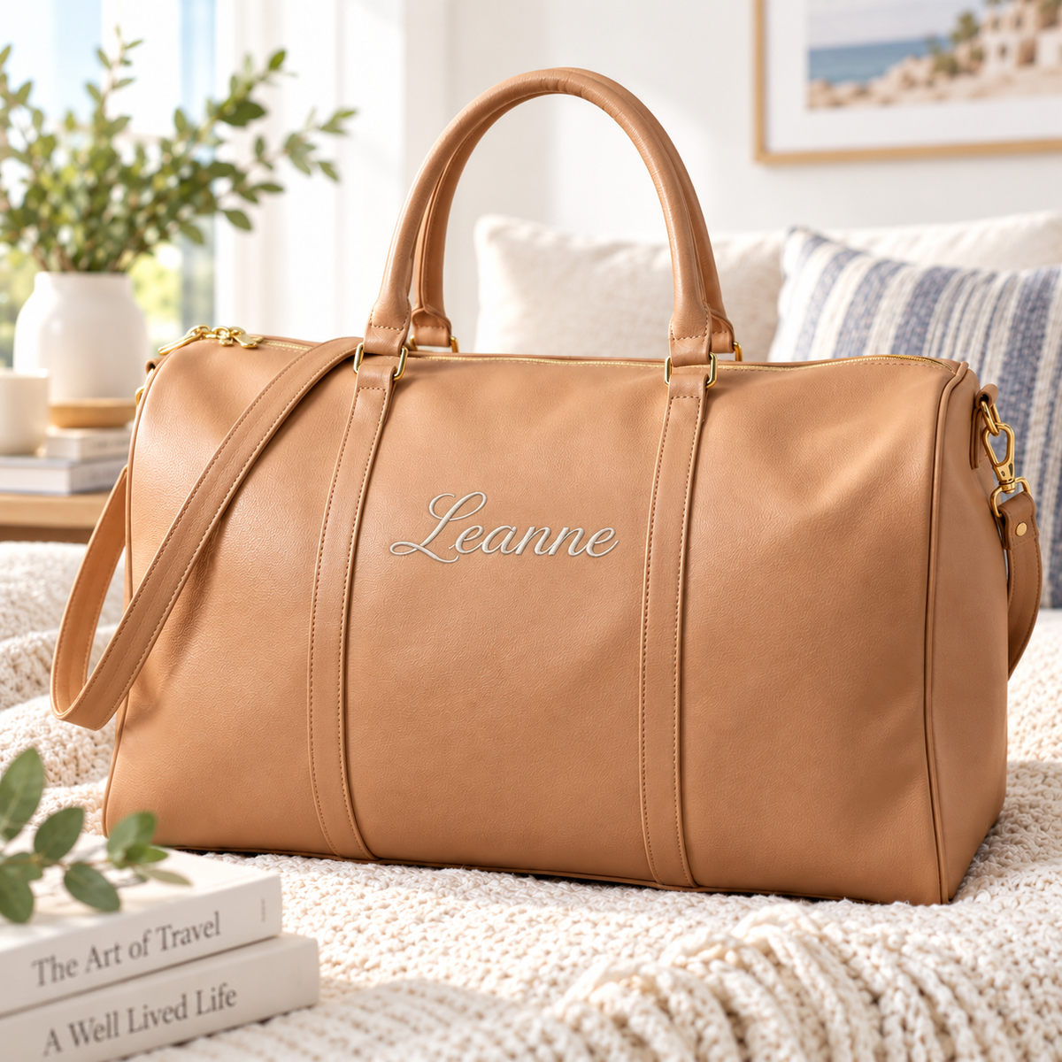 A tan leather duffel bag with gold-tone hardware and the name 'Leanne' embroidered in white script on the front, styled on a cream knit blanket in a bright living room setting.