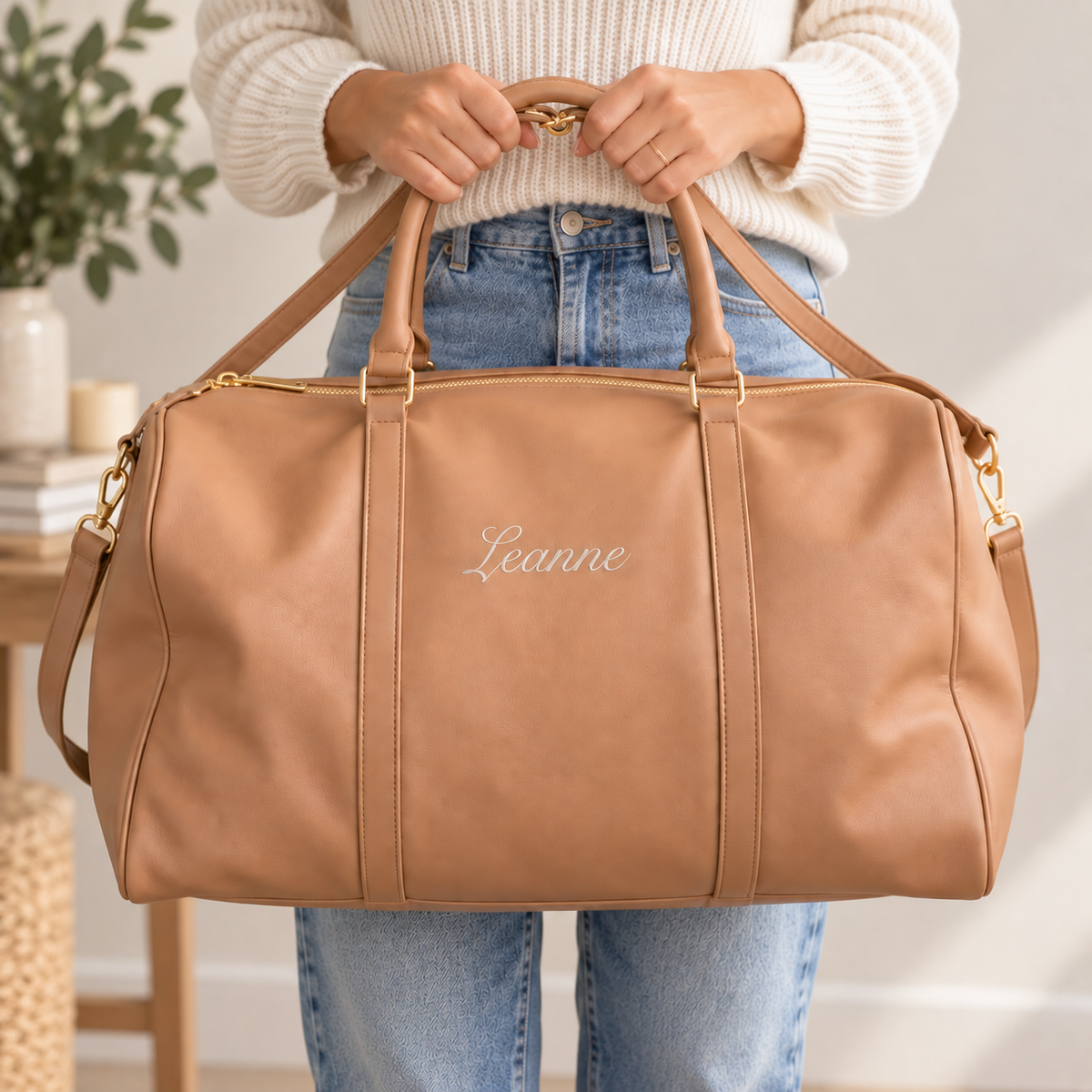 A person in a cream knit sweater and blue jeans holds a tan leather duffel bag with gold-tone hardware and the name 'Leanne' embroidered on the front, photographed in a bright indoor setting.