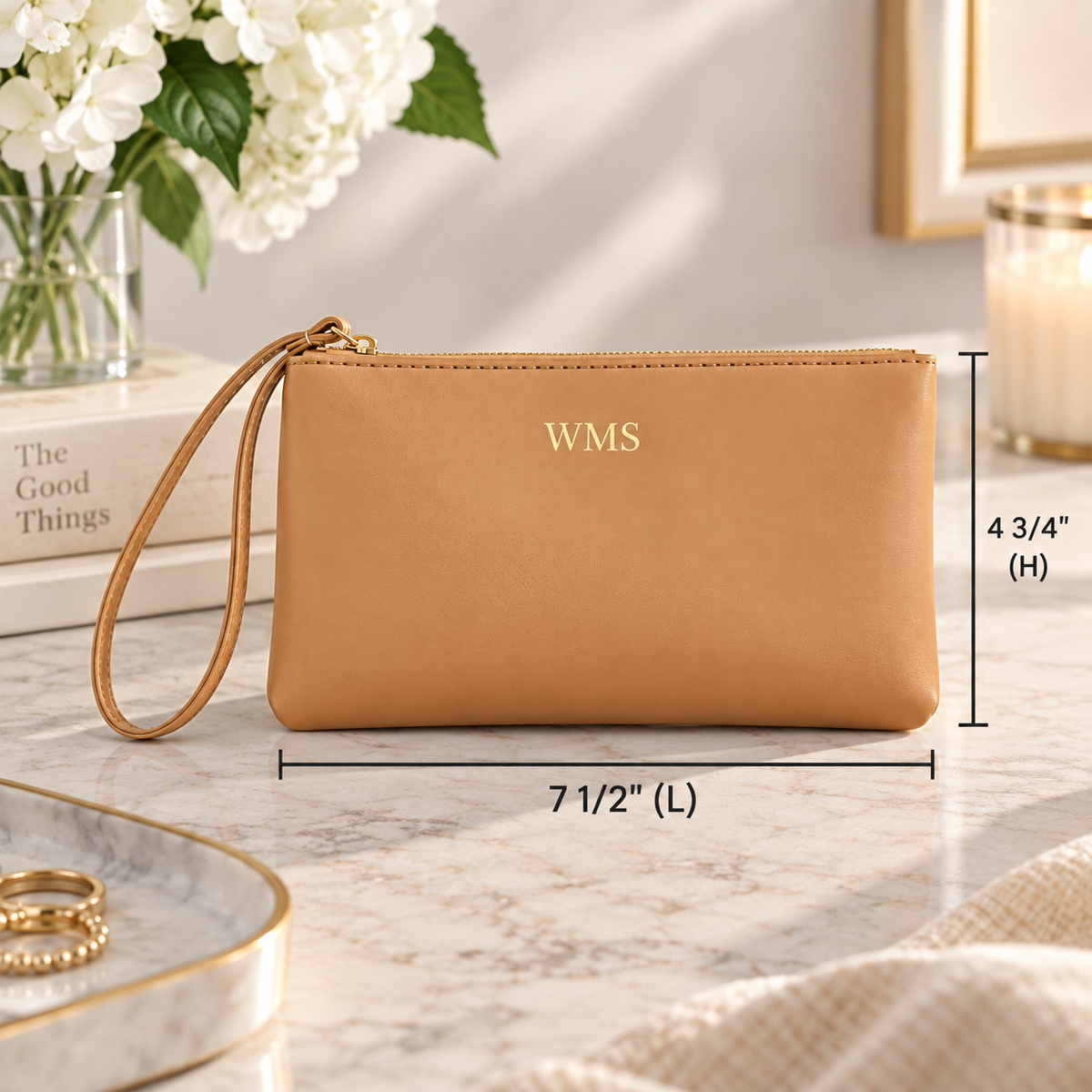 A tan leather wristlet clutch with a wrist strap and gold zipper, shown on a marble surface with dimension lines indicating 7 1/2 inches by 4 3/4 inches. The front is embossed with the initials 'WMS'.