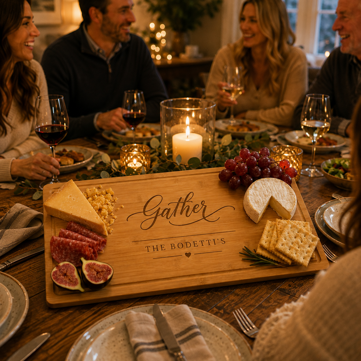 Friends smiling around a candlelit dinner table with wine and a personalized bamboo charcuterie board engraved 'Gather' and 'The Bodetti's' holding cheese, salami, grapes and figs.