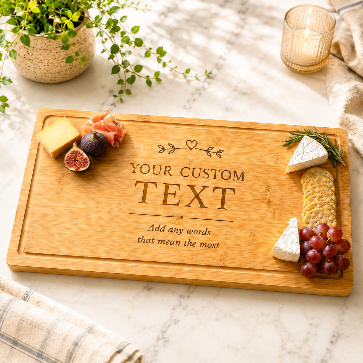 A rectangular wooden charcuterie board with engraved customizable text, styled on a white marble surface with cheese, crackers, grapes, figs, rosemary, and a potted plant.