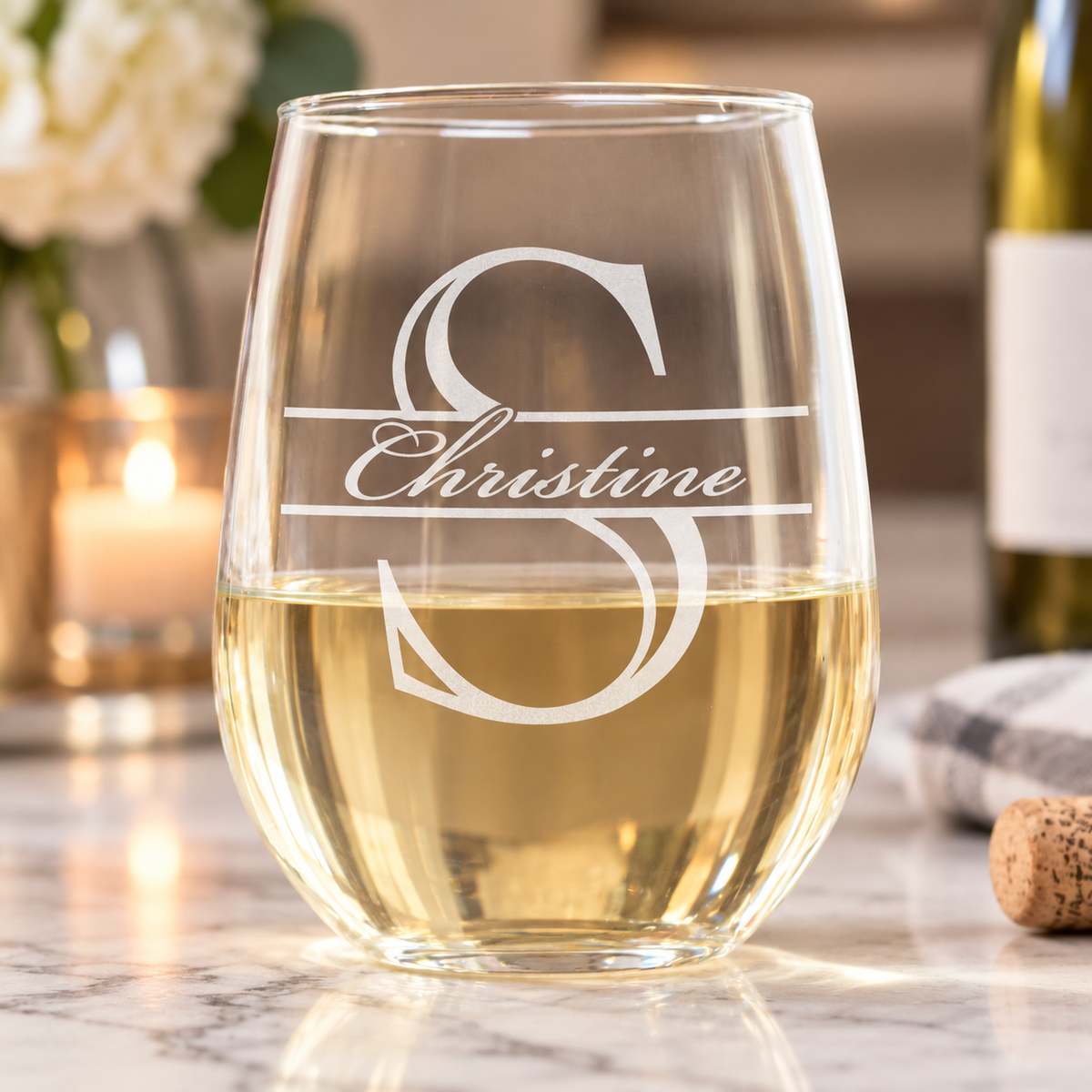 Stemless wine glass engraved with a monogram letter S and the name Christine, filled with white wine on a marble counter with a lit candle and flowers nearby.