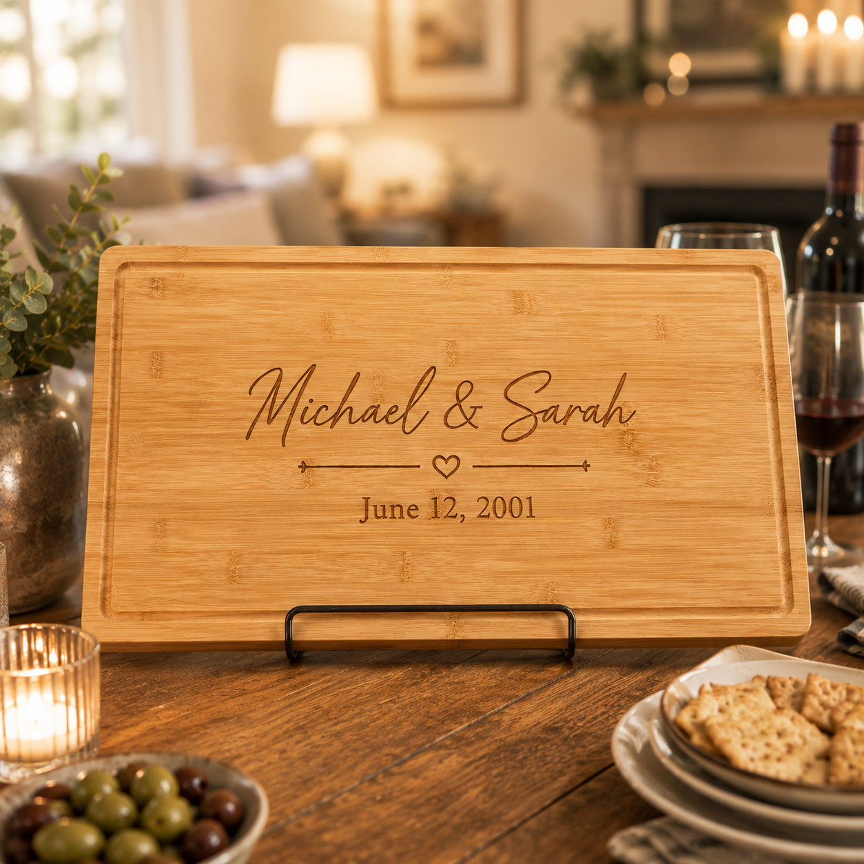 A personalized wooden serving board on a black metal stand, engraved with the names 'Michael & Sarah' and the date 'June 12, 2001', displayed on a wooden dining table with candles, crackers, and wine glasses in a softly blurred living room background.